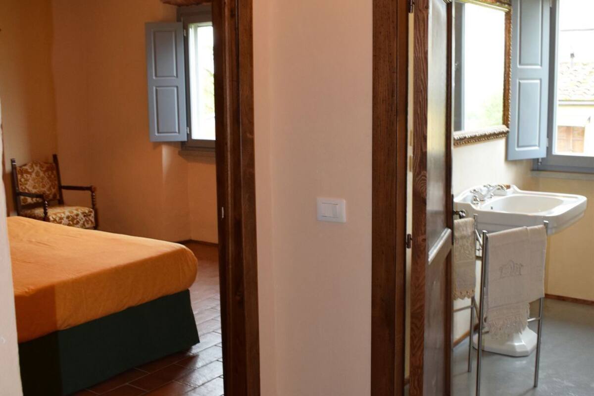 Ein Schlafzimmer mit Bett, Stuhl und Fenster. Es gibt ein Badezimmer mit Waschbecken und Spiegel.