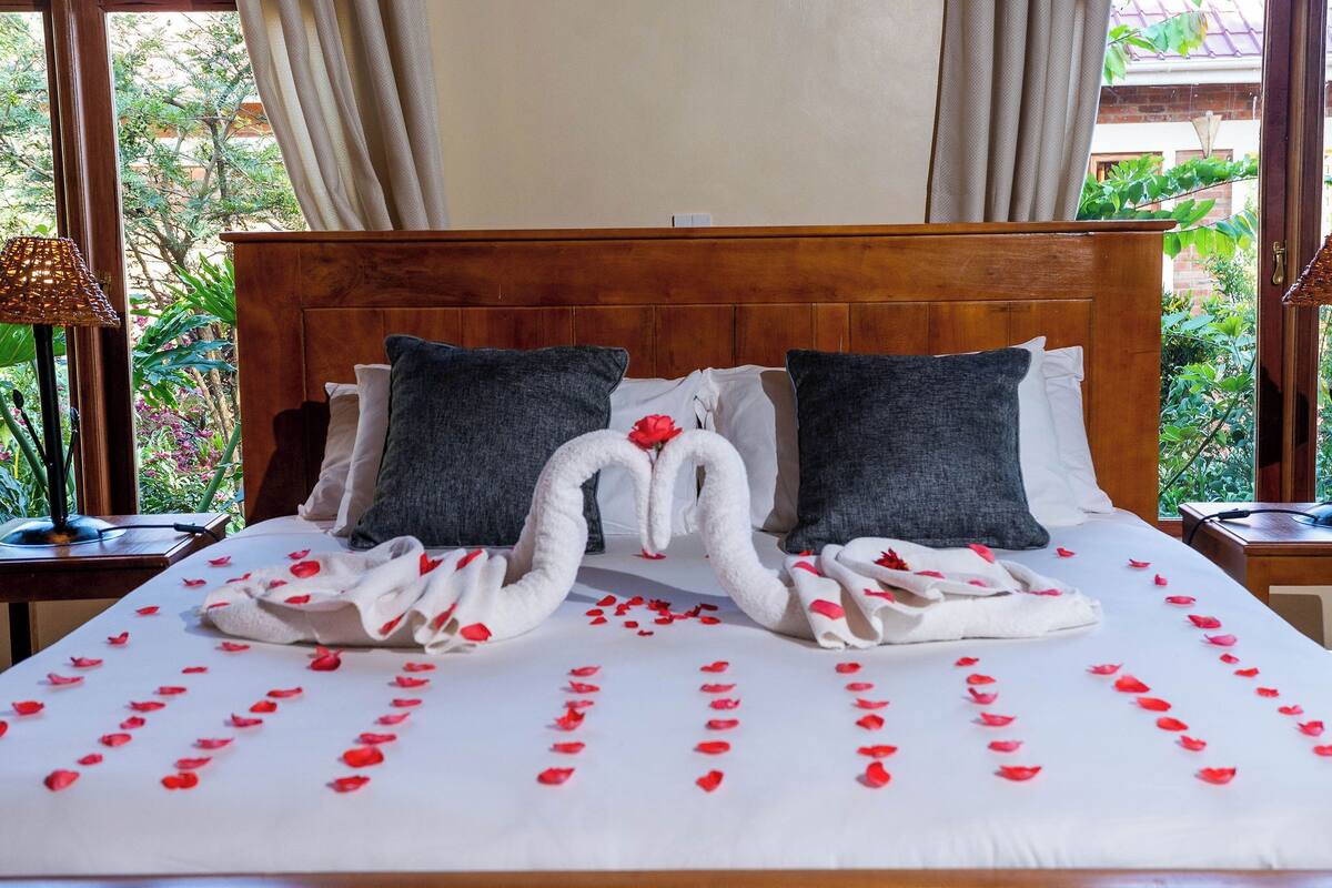 A neatly made bed with a heart-shaped towel arrangement and rose petals, a wooden headboard, and a view of greenery through the window.
