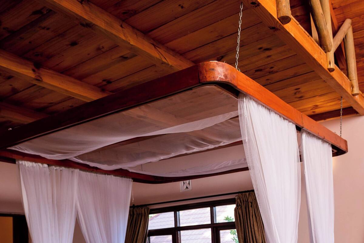 A bedroom with a four-poster bed, wooden ceiling beams, and a view of greenery outside.