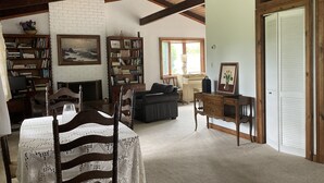 Amplia sala de estar con chimenea, estantería, comedor con mesa y sillas, y un ventanal grande con cortinas.