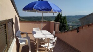 En terrasse med et bord og stole, en blå parasol og udsigt til bakkerne.