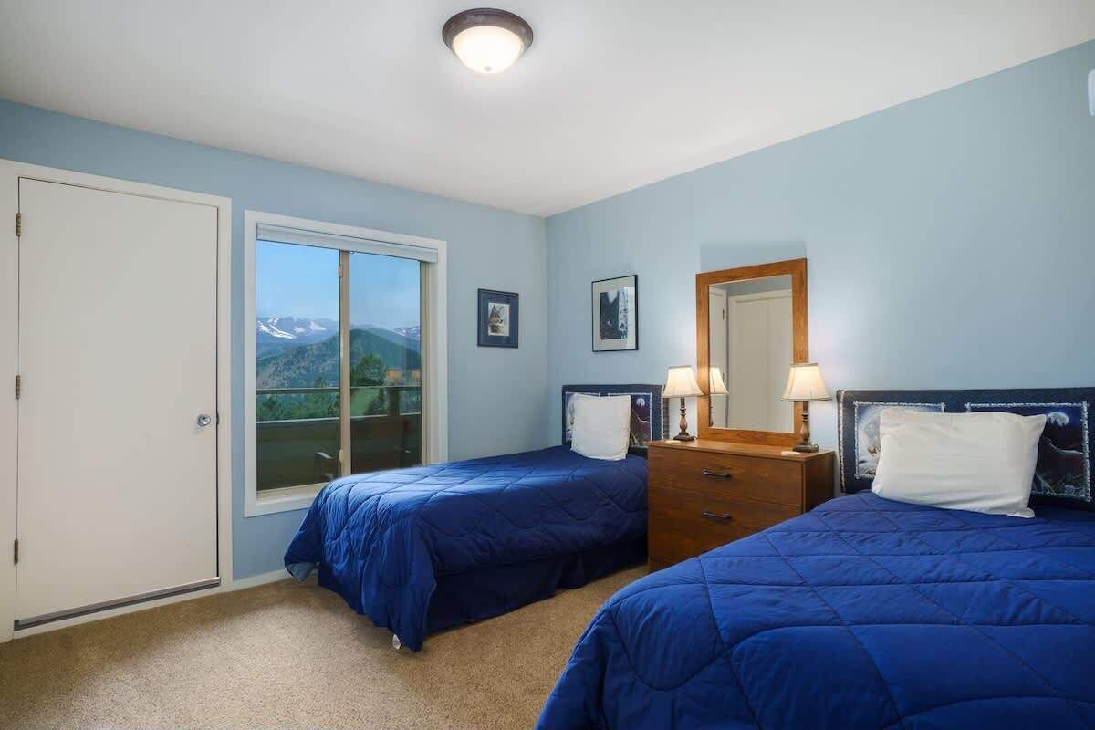 A bedroom with two beds, a dresser, a mirror, and a door leading to a balcony with a mountain view.