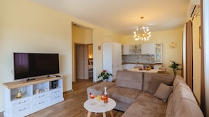 A modern living room with a large flat-screen TV, a white entertainment unit, a beige sofa, and a small round coffee table with drinks and a bottle. - Holiday Home Sea - Two Bedroom Beach House (Petrovac)
