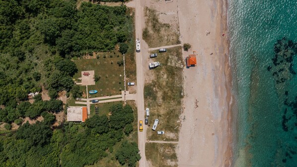 Vista aérea de una zona costera con una playa, vehículos estacionados y un pequeño edificio de techo rojo.