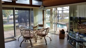Une pièce spacieuse avec une table et des chaises en verre, un téléviseur, et une vue sur une piscine et un espace extérieur.
