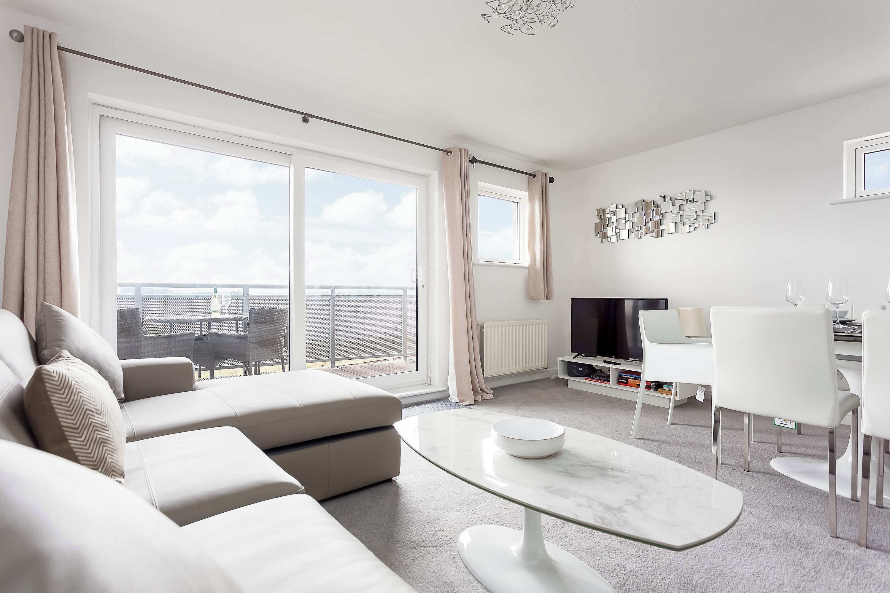 Ein modernes Wohnzimmer mit einer Couch, einem Glastisch, einem Essbereich mit Stühlen, einem Fernseher und Blick auf die Stadt durch große Fenster.