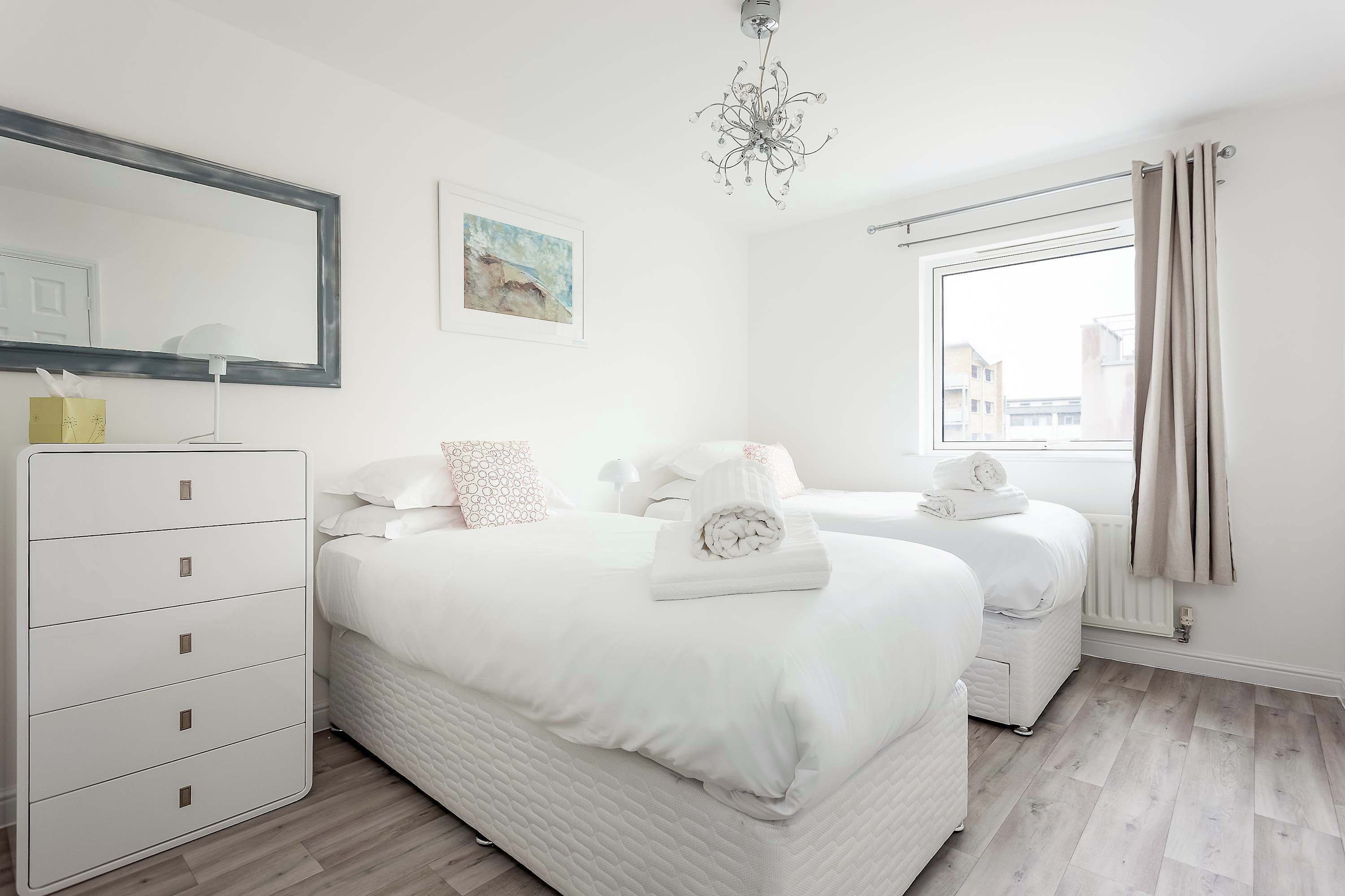 A modern bedroom with two beds, a dresser, a chandelier, and a window with curtains.