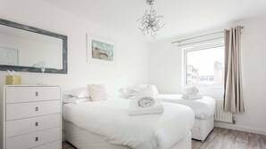 A modern bedroom with two beds, a dresser, a chandelier, and a window with curtains. - Beach Point - Stunning Harbour Views from Balcony (Poole)