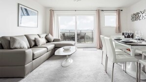 A modern living room with a grey sectional sofa, a round coffee table, and a dining area with white chairs and a glass table. - Beach Point - Stunning Harbour Views from Balcony (Poole)