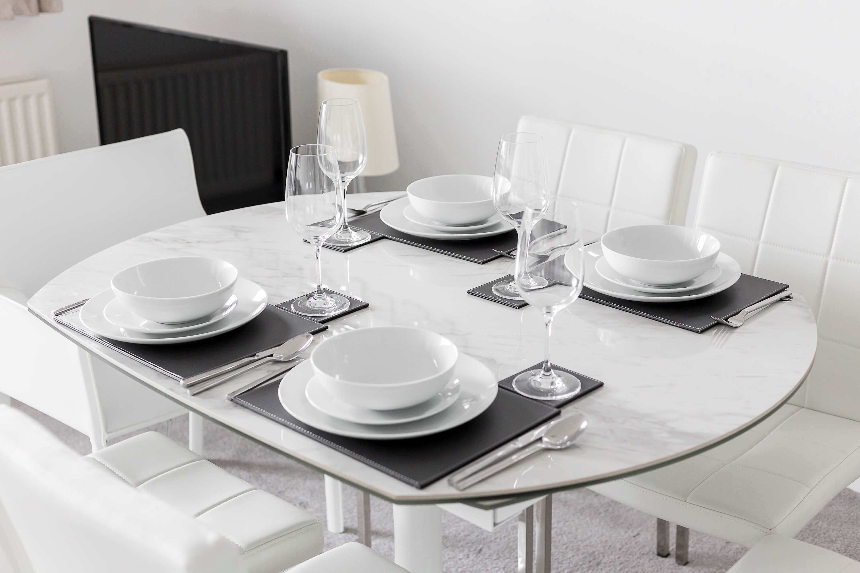 A dining table set with white plates, glasses, and cutlery, surrounded by white chairs and a black cushioned bench.