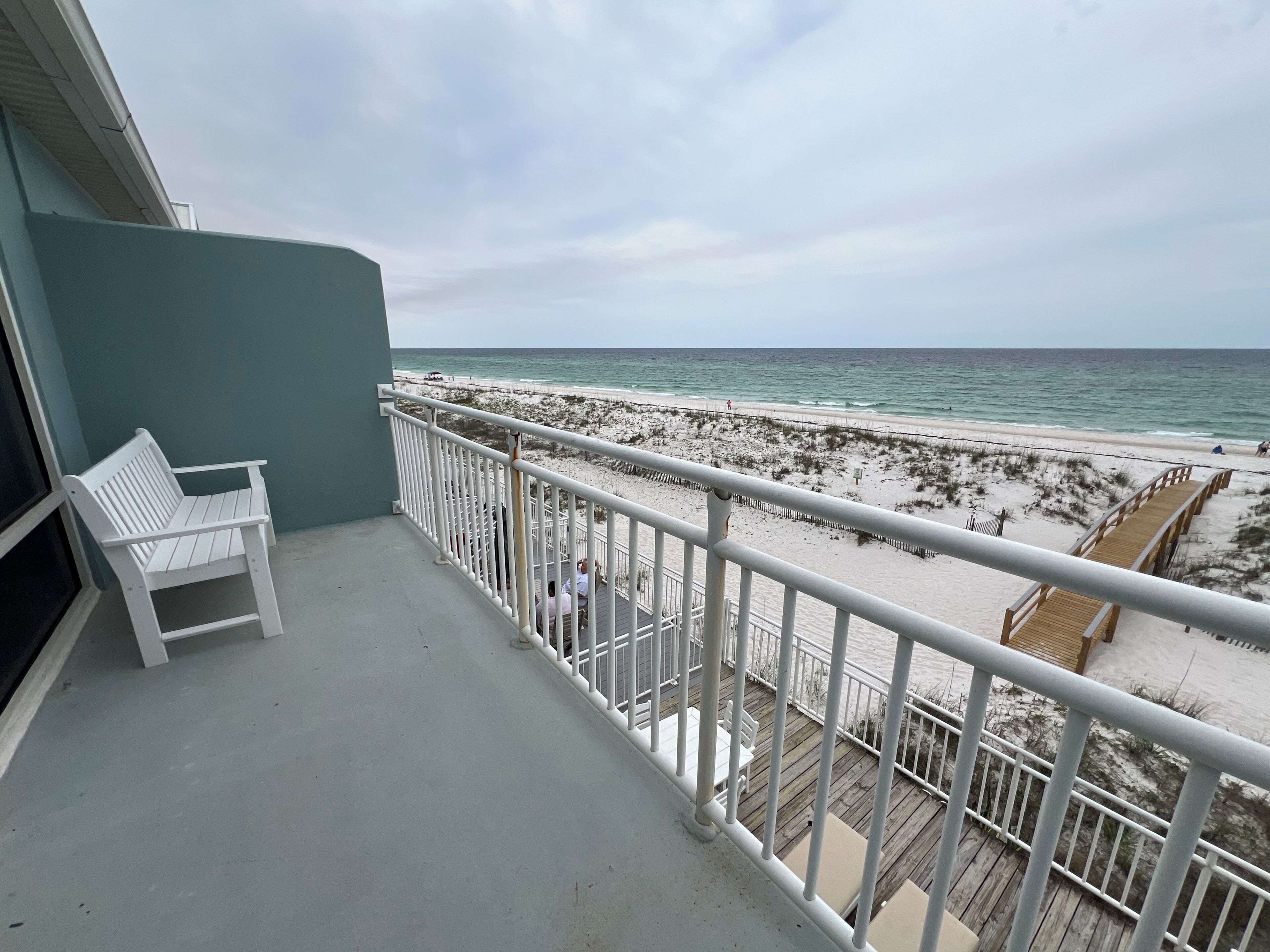 Un balcon avec une chaise blanche, une terrasse en bois et une vue sur une plage avec une promenade en bois.