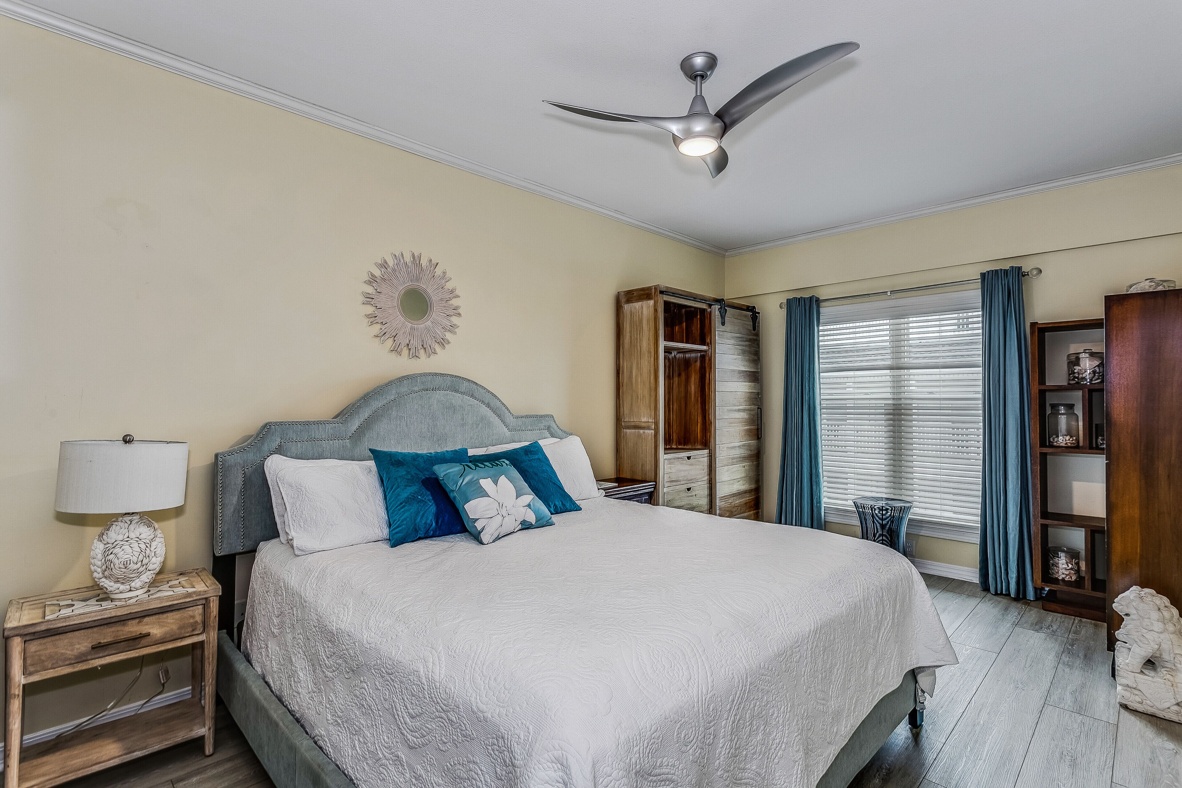 A neatly arranged bedroom with a bed, nightstand, lamp, ceiling fan, and a window with curtains.