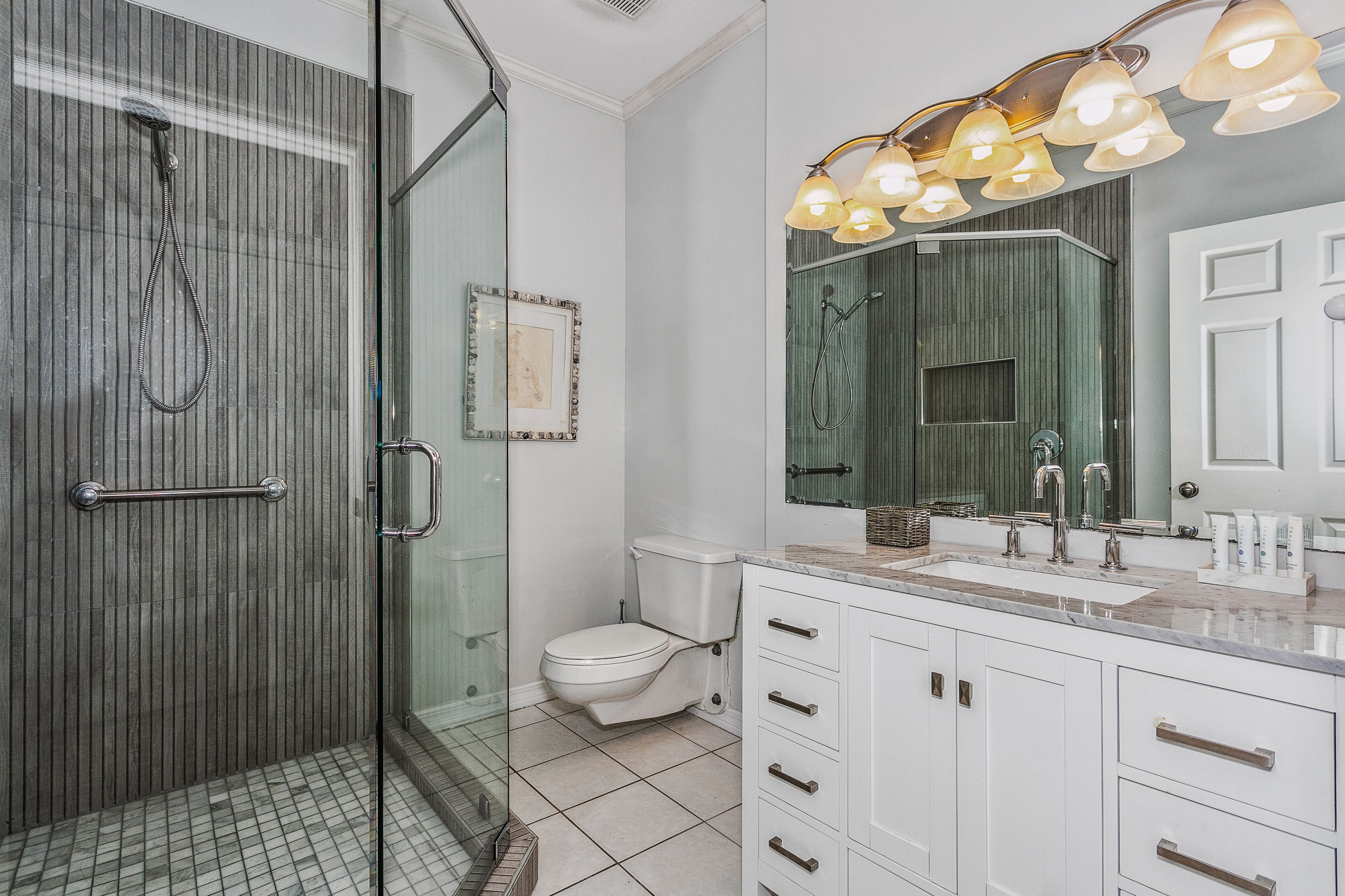 A modern bathroom with a glass-enclosed shower, a white toilet, and a vanity with a large mirror and multiple light fixtures.