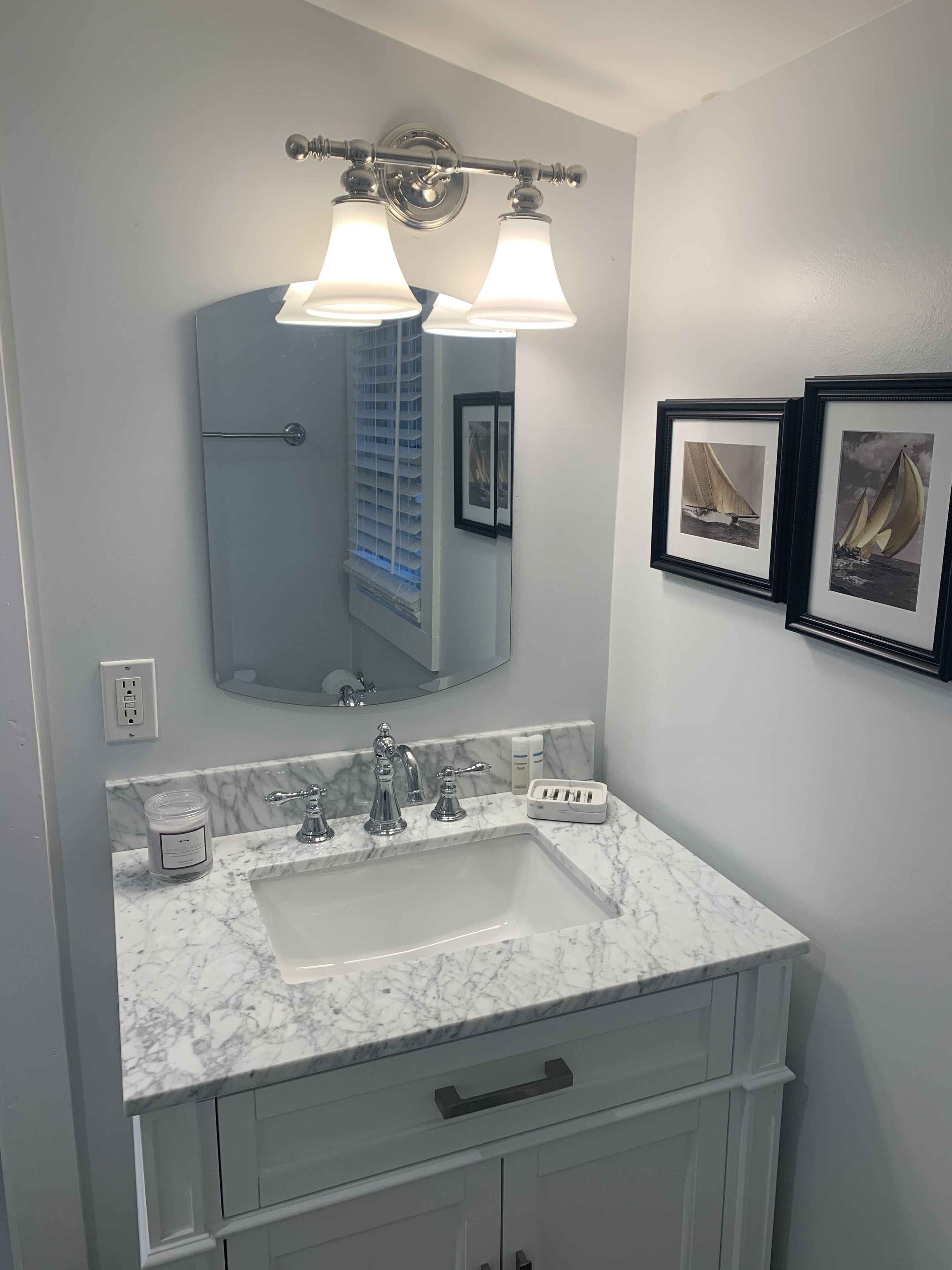 A bathroom with a large mirror, two wall-mounted lights, a marble sink, and a framed picture of a sailboat.