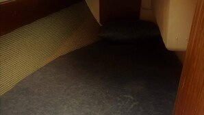 A compact bed with a grey mattress in a small room with a wooden bench and a shelf with various items.