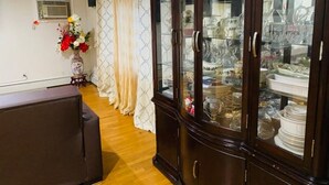 Een kamer met een houten kastje met servies en glazen, een tafeltje met een bloevas, en een raam met gordijnen.