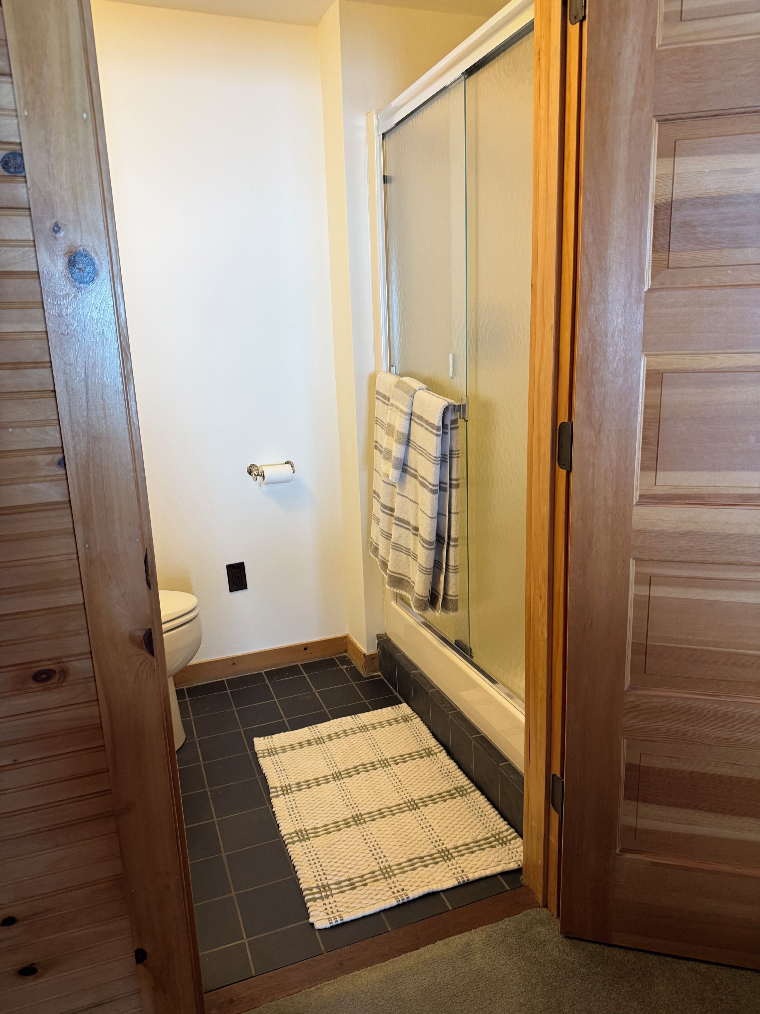 Un baño con inodoro, ducha con puerta de cristal, toalla colgada en la pared y alfombra estampada en el suelo.