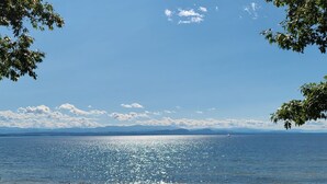 A beach with clear water, a distant sailboat, and mountains in the background. - Designer Lake Champlain Retreat with Adirondack Views & Private Waterfront (Willsboro)
