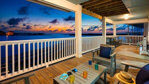Eine überdachte Terrasse im Freien mit Holzboden, weißen Geländern und Blick auf das Meer bei Sonnenuntergang.