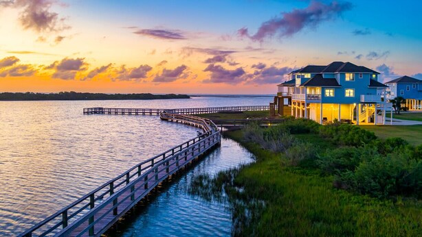Ein Anwesen am Wasser mit einem hölzernen Steg, einem mehrstöckigen Haus und Blick auf den Sonnenuntergang über dem Meer.