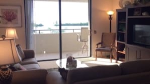 A living room with a sofa, armchair, and a television. There is a balcony with a chair and a lamp. - Hudson Hideaway: 2 BR/2 Bath Gulf Front Condo,Stunning Sunset from 27' Balcony (Hudson)