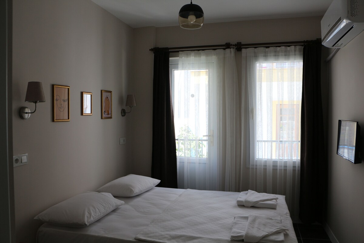 A hotel room with a bed, a window with curtains, wall-mounted lights, and framed pictures.