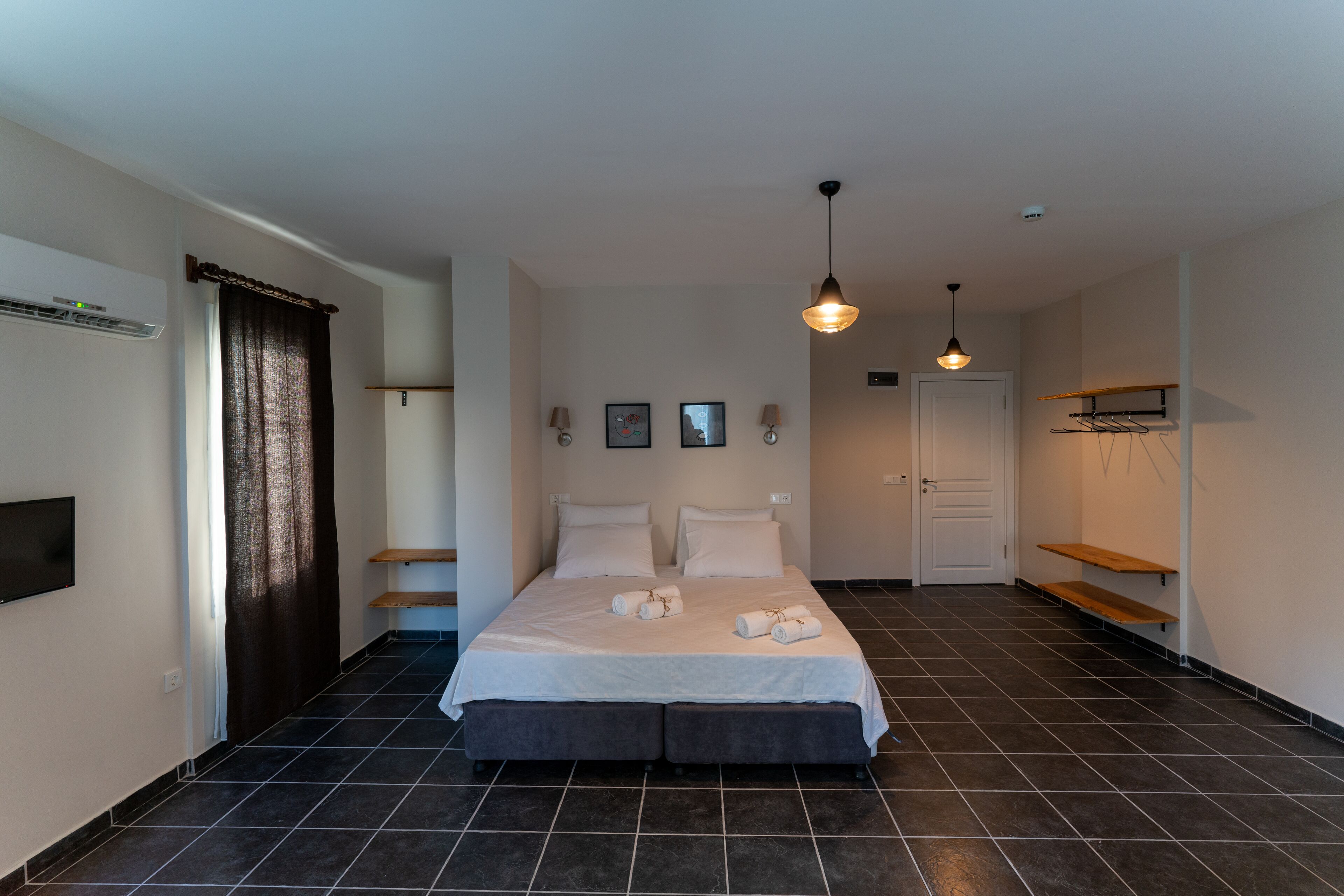 A bedroom with a large bed, two bedside tables, a wardrobe, a hanging light fixture, and a flat-screen TV mounted on the wall.