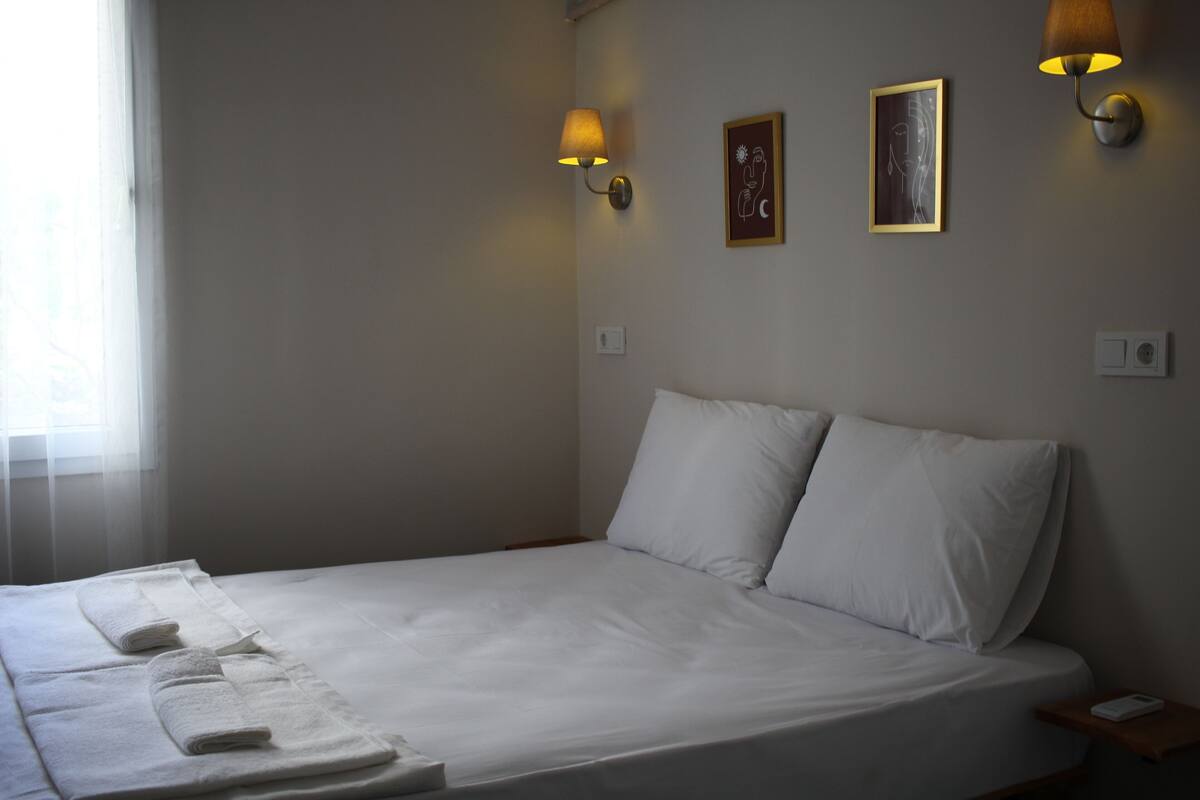 A neatly made bed with white linens and two pillows, a bedside table with a lamp, and two wall-mounted light fixtures.