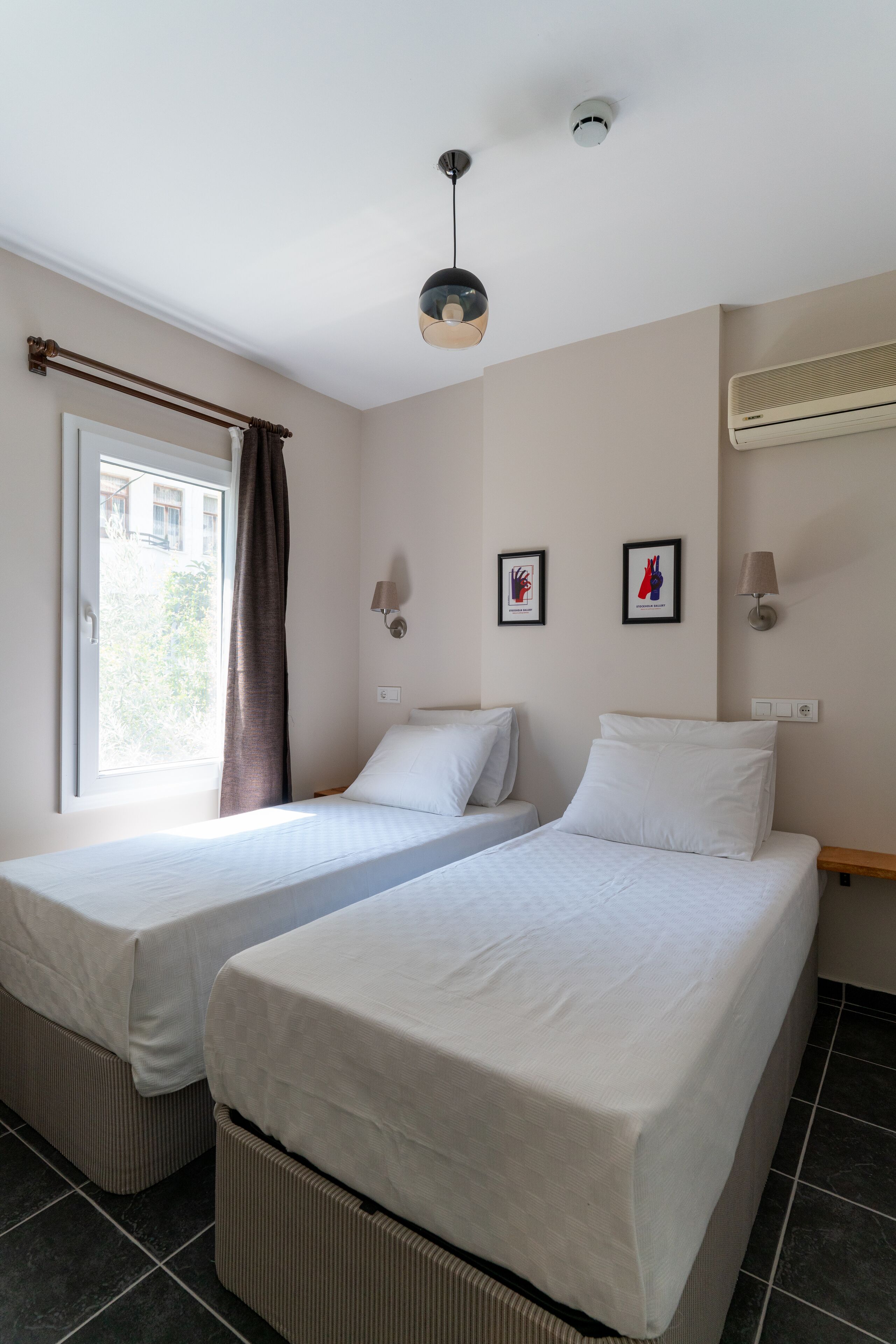 Una habitación de hotel con dos camas individuales, una ventana con cortinas, una lámpara en el techo, aire acondicionado instalado en la pared y cuadros enmarcados.