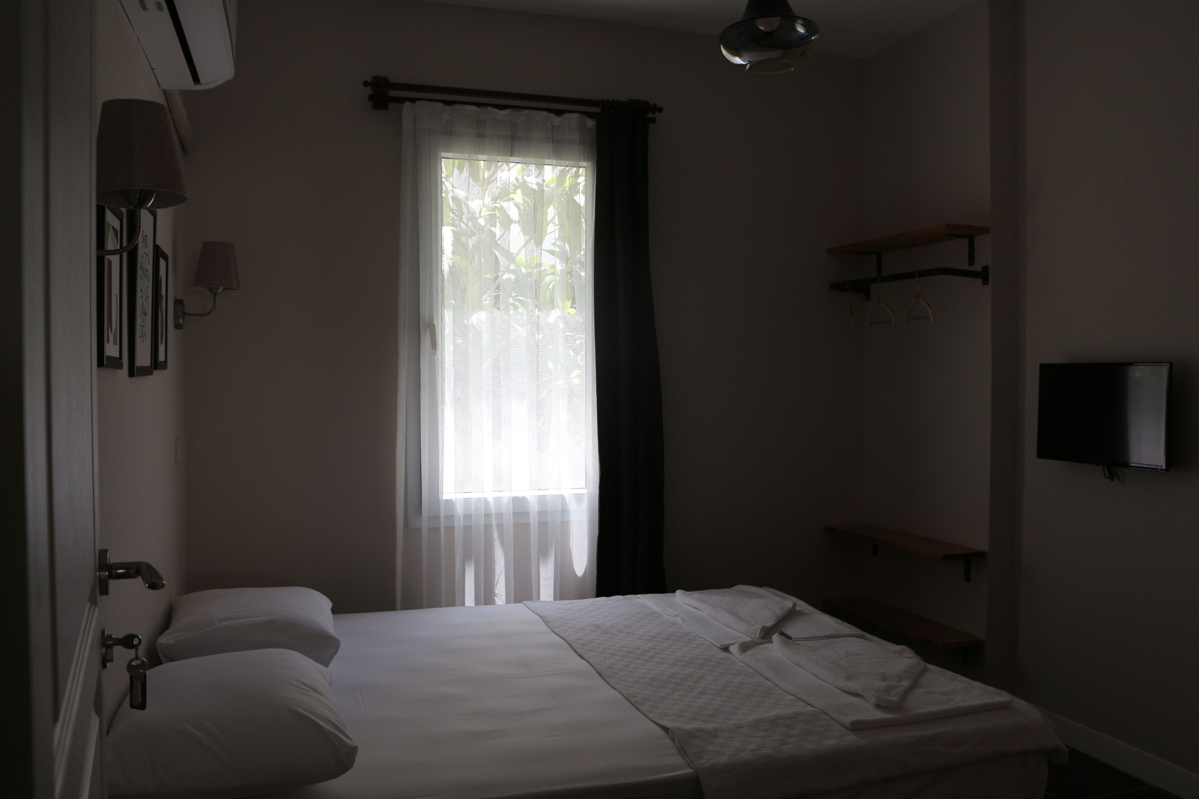 A bedroom with a bed, a window with curtains, a wall-mounted TV, and a bedside table.