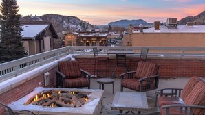 Eine Dachterrasse mit Sitzgelegenheiten im Freien, einer Feuerstelle und Blick auf eine Stadt mit Gebäuden und Bergen im Hintergrund.