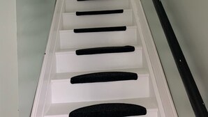 Un escalier avec des marches blanches et des rampes noires, menant à un palier où est accrochée une image encadrée au mur.