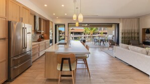 A modern kitchen with a large island, wooden cabinets, and stainless steel appliances. There is a dining area with a table and chairs, and a living room with a sofa and a TV. - KLVR welcomes you to Honua Kai Luana 11C Oceanfront Resort Resort and POOL view  (Lahaina)