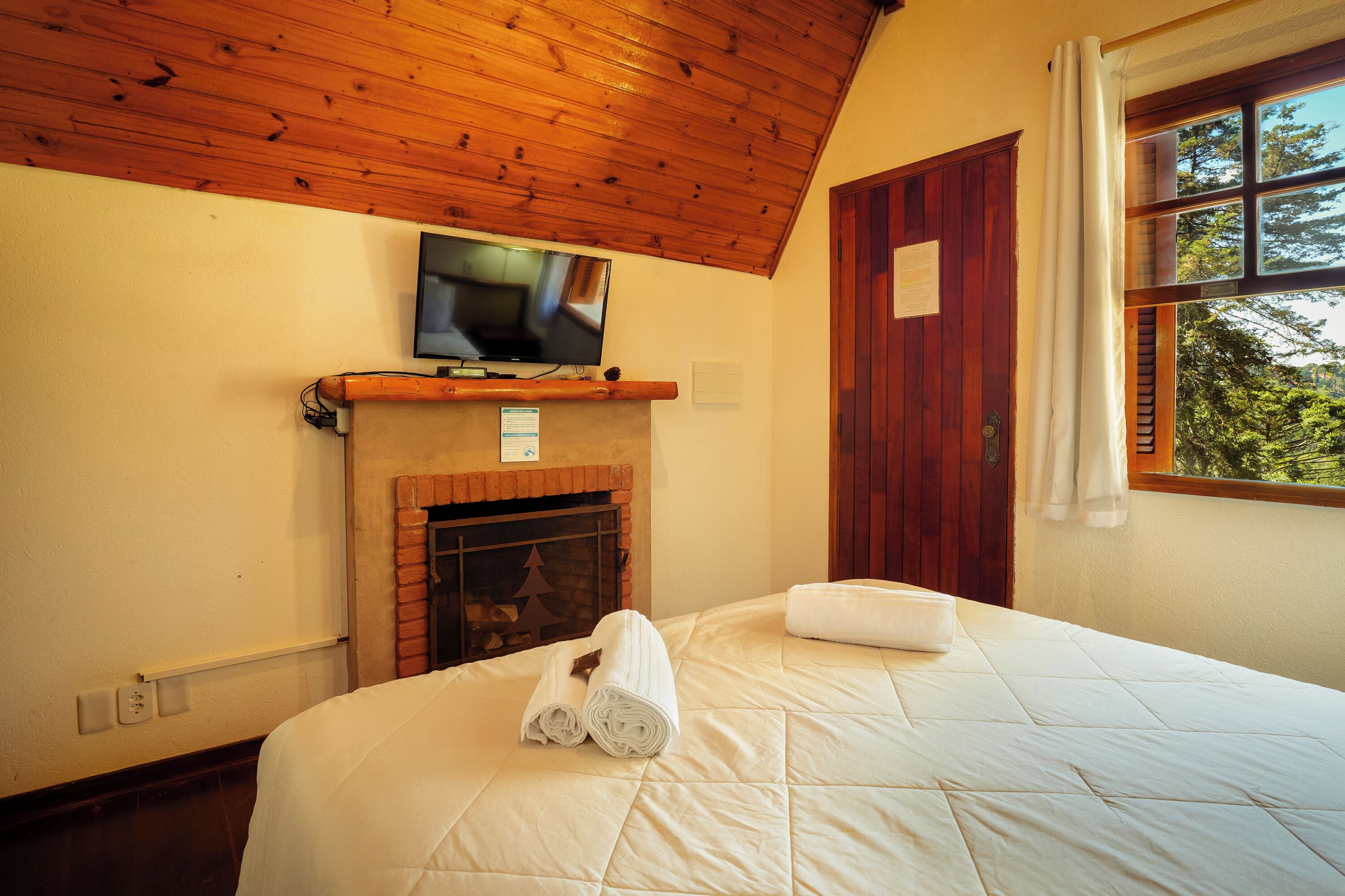 Habitación de hotel con techo de madera, televisor sobre la chimenea, cama con colcha y ventana con cortinas.
