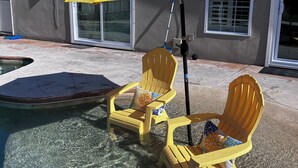 Un espace aménagé au bord de la piscine, avec deux chaises Adirondack jaunes et un parasol jaune, une piscine à l’eau claire, et une maison dotée de grandes fenêtres et d’une porte.