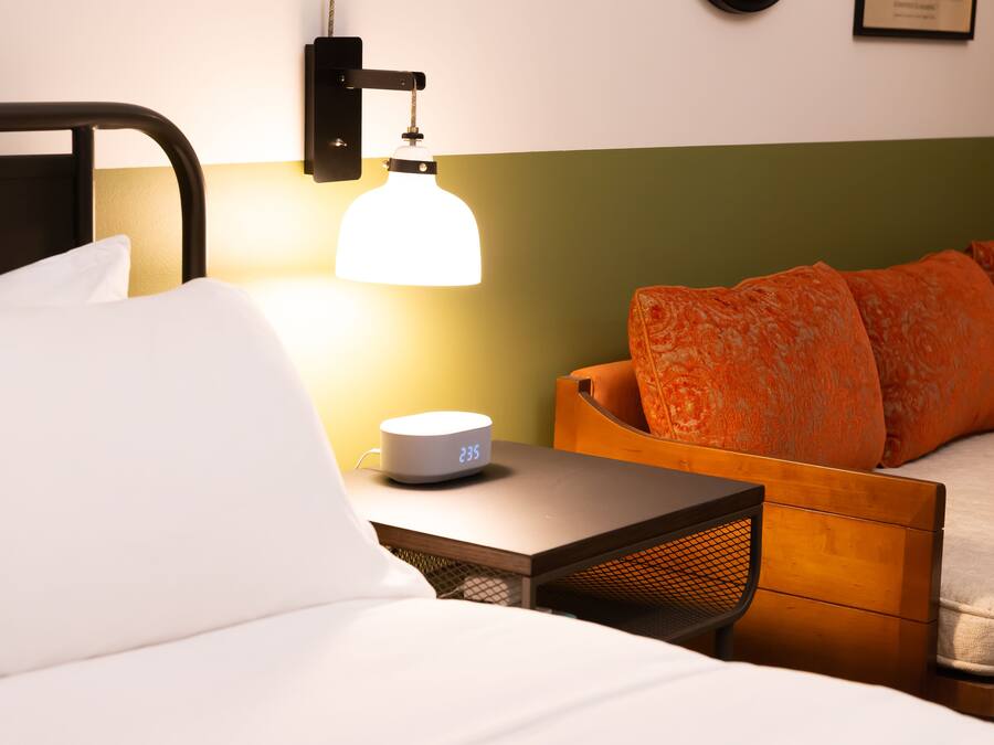 Una cama bien hecha con sábanas blancas, una mesita de noche con un reloj digital y una lámpara de pared.