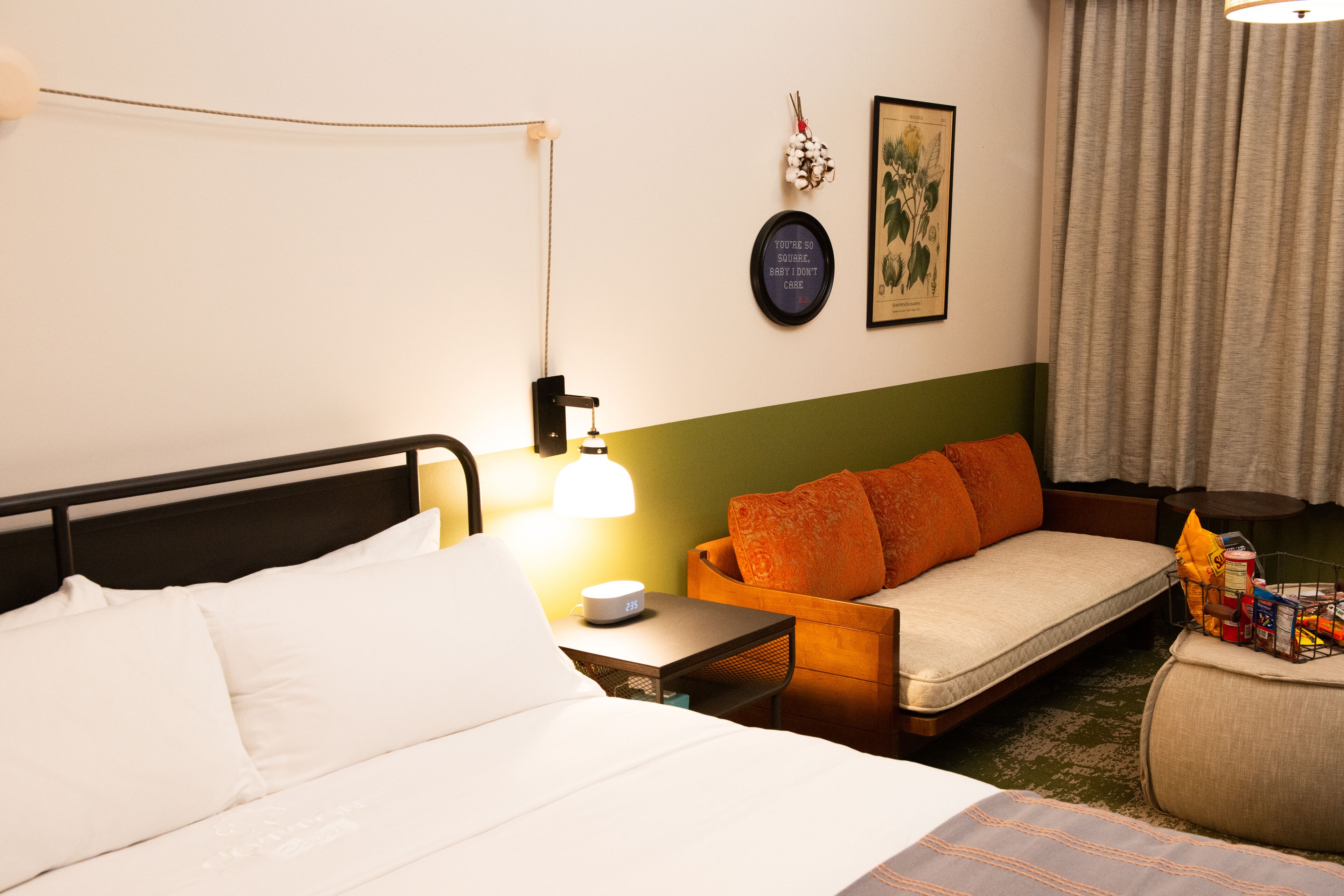 A hotel room with a bed, a sofa, a small table with a lamp, and a wall with a framed quote and a botanical illustration.