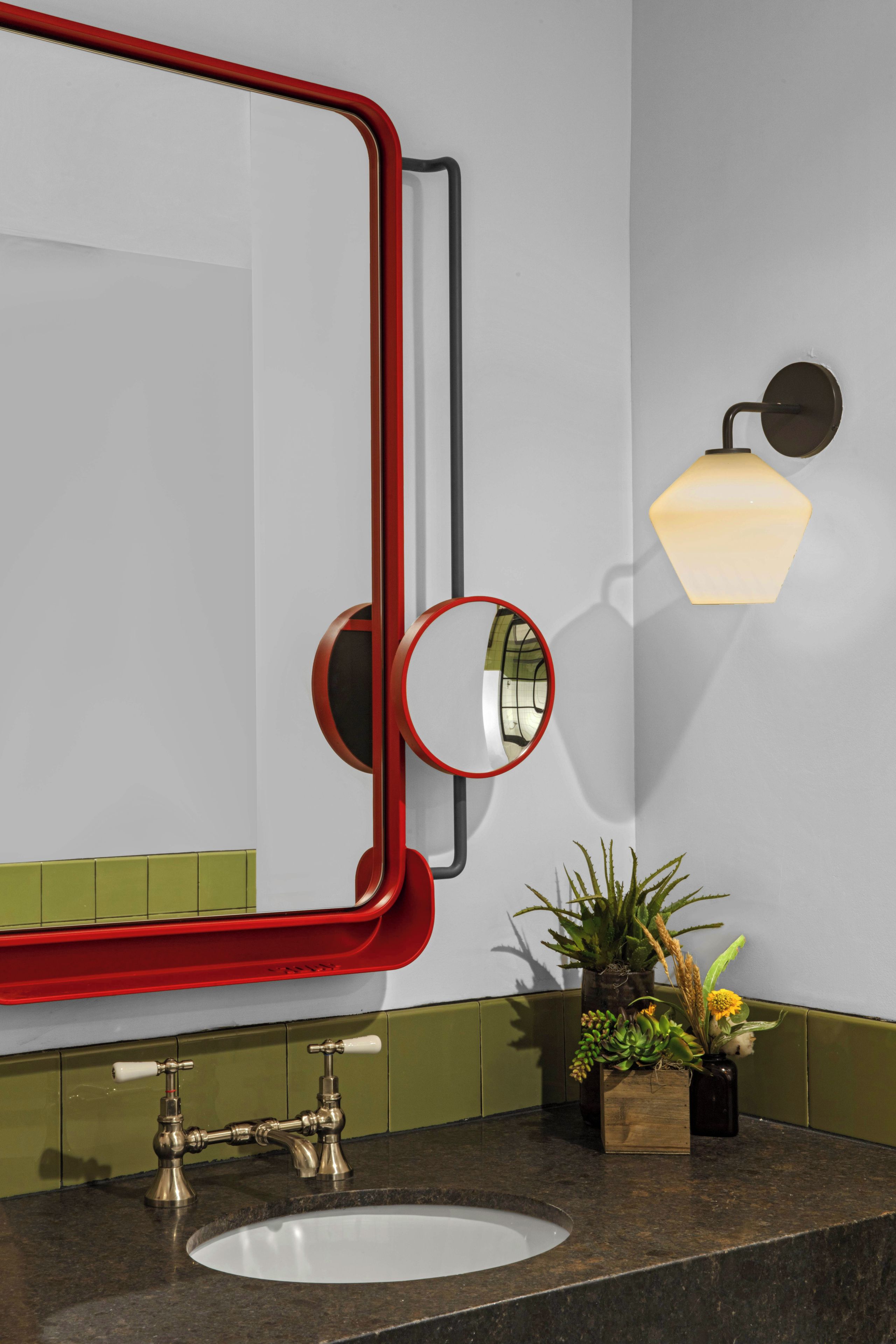 A bathroom with a white sink, a red-framed mirror, a wall-mounted light fixture, and a potted plant.