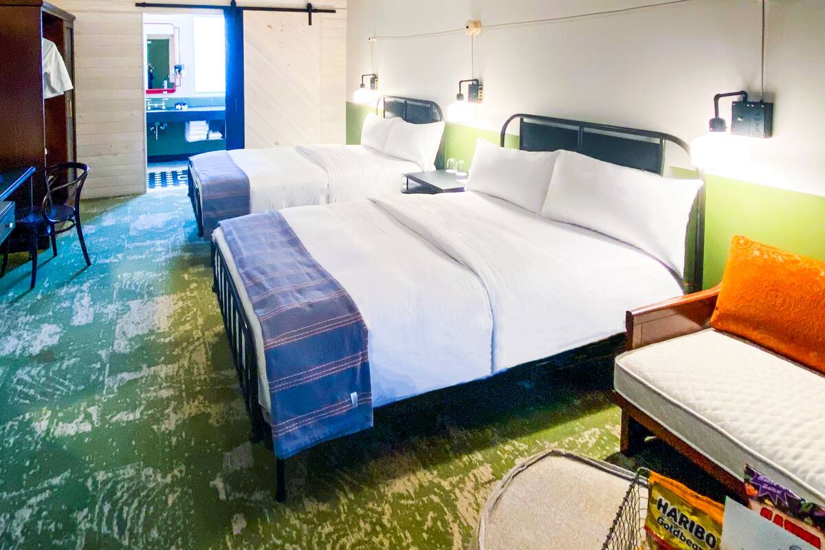 Ein Hotelzimmer mit zwei Betten, einem Schreibtisch, einem Sessel und einem Deckenventilator, sowie einem großen Fenster.