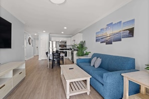 A modern living room with a blue sofa, a wooden coffee table, and a TV on the left. A kitchen is visible in the background with white cabinets and a microwave. - Beautiful Premium Brand New 2 Bedroom Condo w/ Ocean View  (Myrtle Beach)
