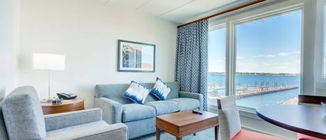 Een moderne hotelkamer met een grijze bank, een houten salontafel en een rode stoel. Een groot raam biedt uitzicht op zee en een pier.