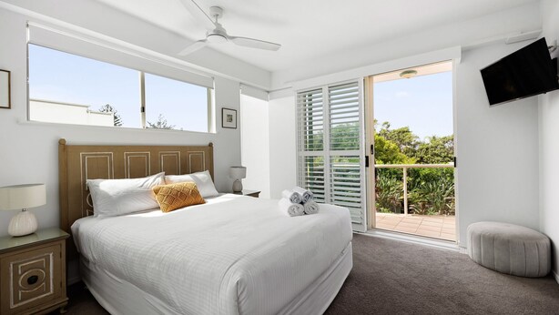 A bedroom with a large bed, a ceiling fan, a TV mounted on the wall, and a balcony with a view of greenery. - Sempre 7 Sunshine Beach (Sunshine Beach)