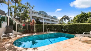 A swimming pool with a tiled deck, surrounded by palm trees and a building with balconies. - Sempre 7 Sunshine Beach (Sunshine Beach)
