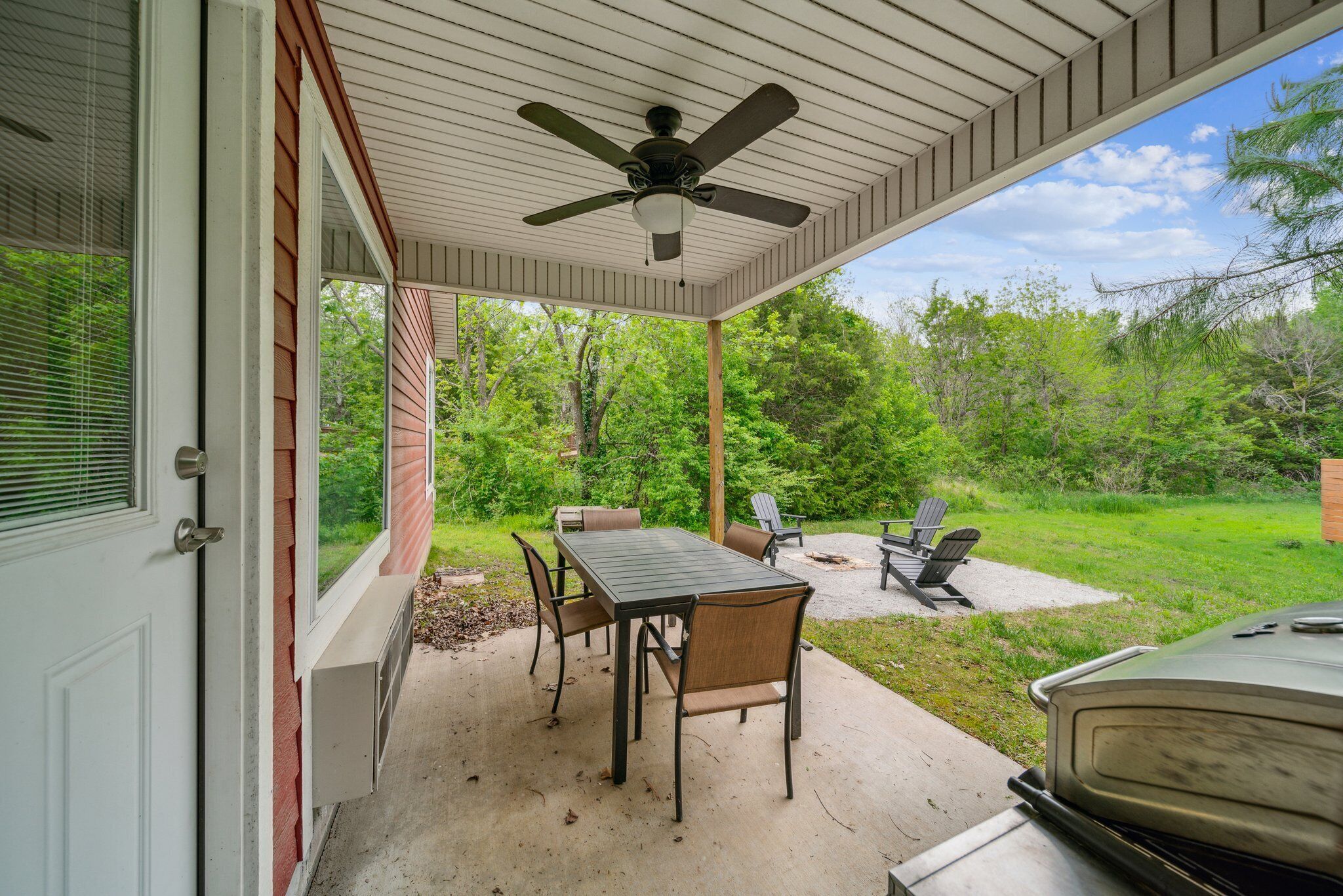A covered patio with a table and chairs, a ceiling fan, and a view of a wooded area.