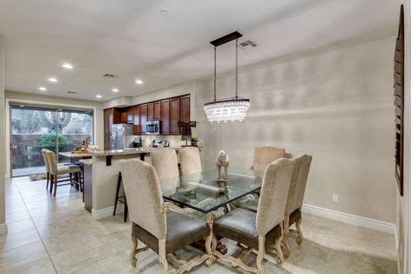 A dining area with a glass table and upholstered chairs, a chandelier, and a kitchen in the background. - La Joya del Desierto (The Jewel of the Desert) - Cave Creek (Cave Creek)