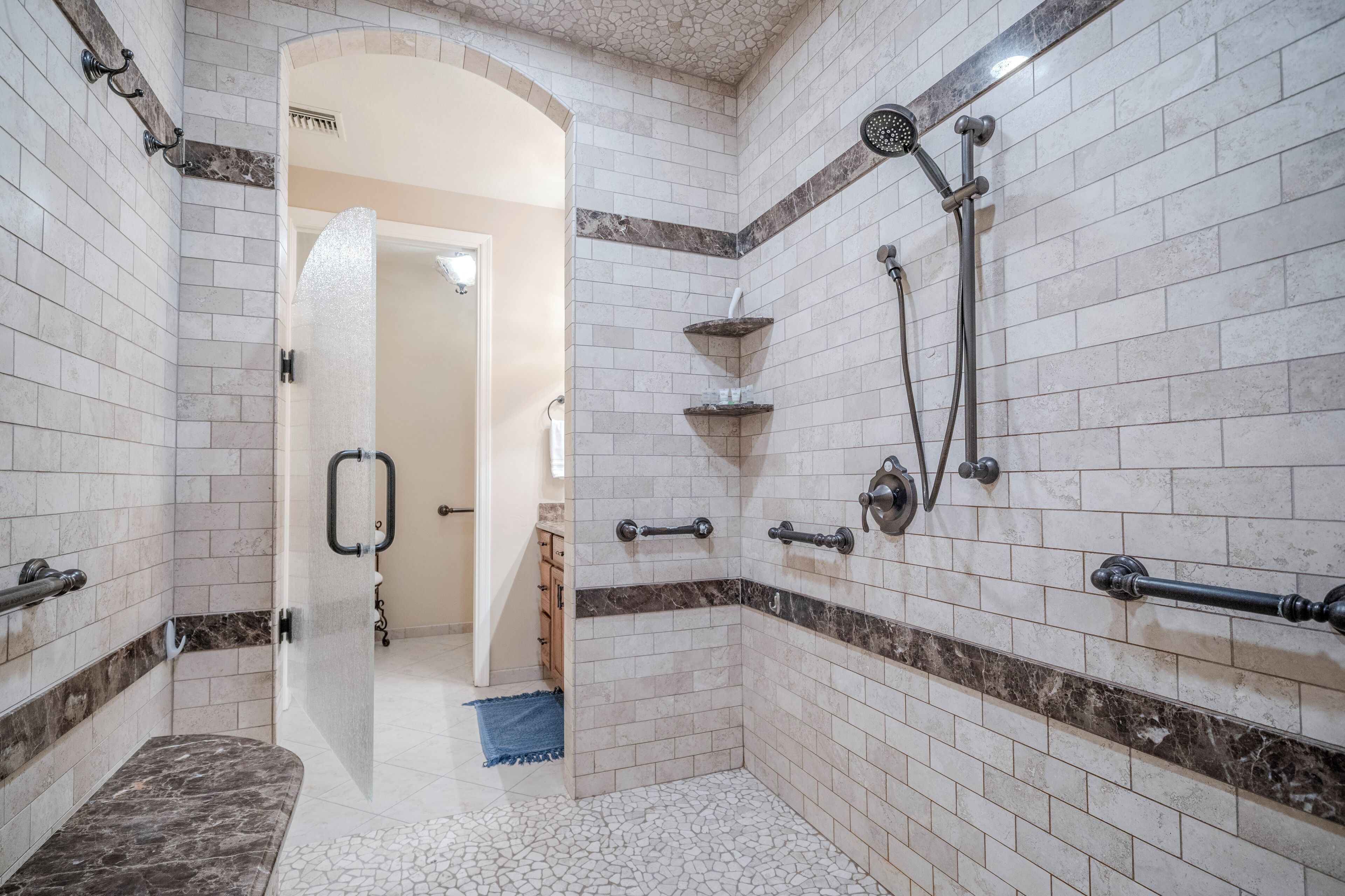 A bathroom with white and brown tiles, a shower with a handheld showerhead, and a bench.