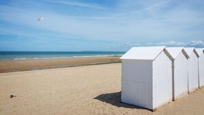 Une rangée de cabanes de plage blanches s’étend le long d’une plage de sable, avec un ciel bleu clair et l’océan en arrière-plan.