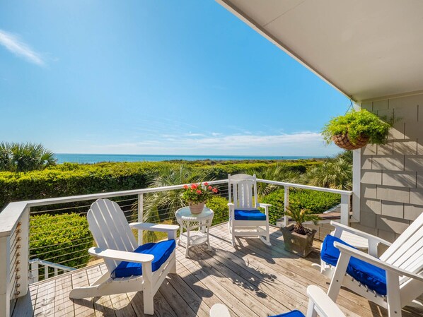 A wooden deck with white chairs and a table, overlooking a coastal landscape.