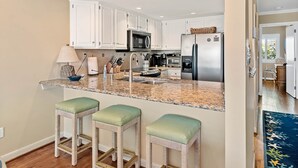 A modern kitchen with white cabinets, stainless steel appliances, and a granite countertop. There are three wicker stools with green cushions.