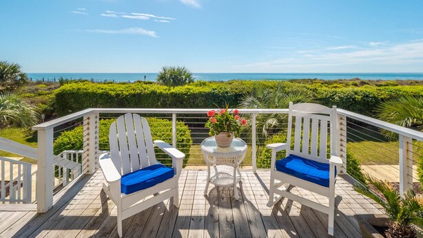A wooden deck with two white Adirondack chairs with blue cushions, a small round table with a glass top, and a potted plant with red flowers.